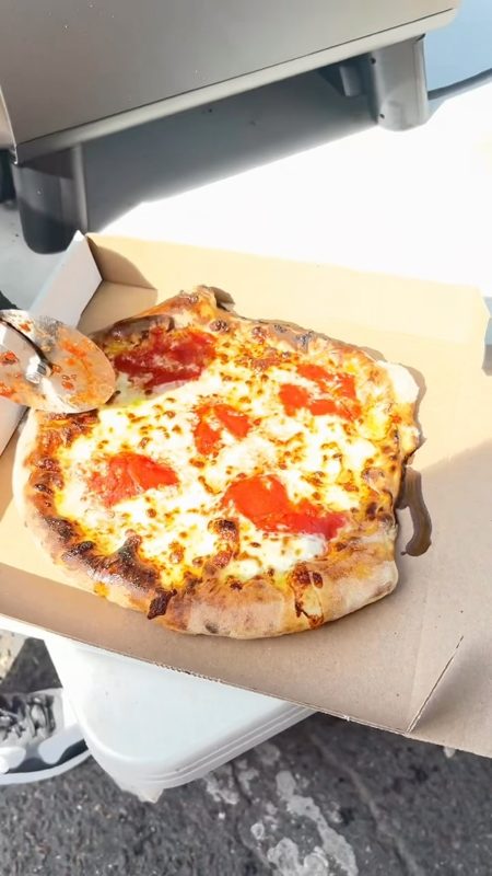 Video still of a cheese pizza with tomato slices being boxed, pizza cutter resting on top